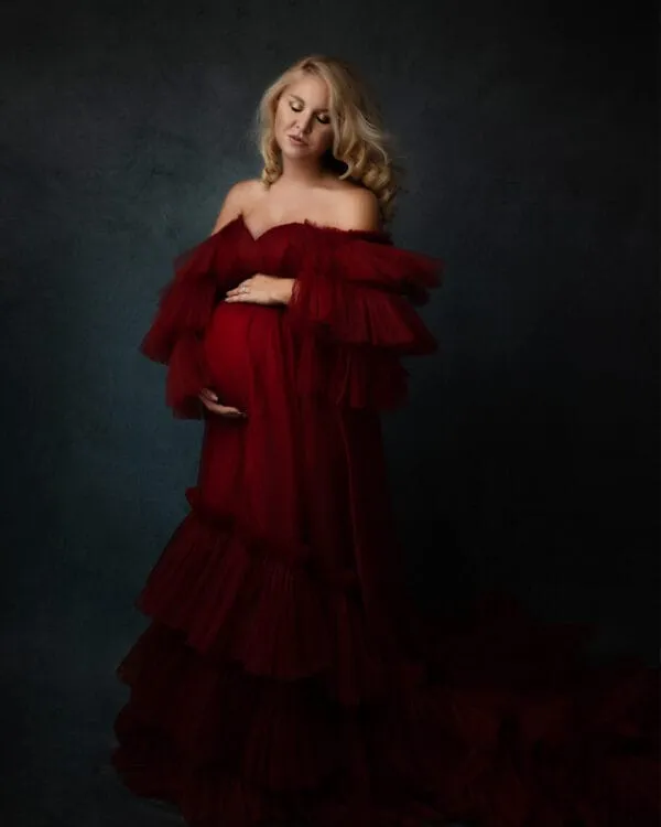 Eine schwangere Frau umhüllt von einem üppigen, schulterfreien roten Kleid, das ihre Formen sanft betont und mit Rüschen zu ihren Füßen fällt.